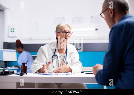 Männliche Rentnerpatientin sitzt gegenüber der pensionierten Ärztin in der Klinik und diskutiert Behandlungspläne. Leitende Ärztin sitzt am Schreibtisch und gibt dem alten weißen Mann im Krankenhauszimmer medizinischen Rat. Stockfoto