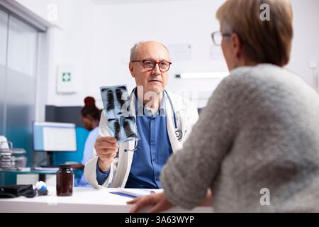 Eine ältere Patientin sitzt gegenüber ihrem pensionierten männlichen Arzt, der in der Krankenhausniederlassung Röntgenuntersuchungen durchführt. Älterer Arzt diskutiert medizinische Befunde mit alter Frau und skizziert Behandlungsmöglichkeiten. Stockfoto