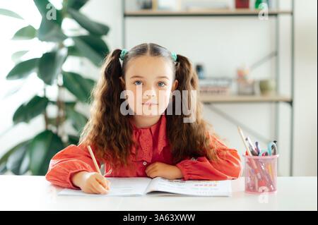 Wunderschönes weißes blauäugiges Schulmädchen 6-7 Jahre in roter Bluse mit lockigen Haaren sitzt zu Hause an einem Schreibtisch, hält einen Bleistift und schaut während der Hausaufgaben in die Kamera Stockfoto