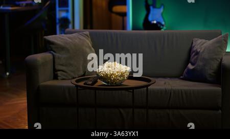 Popcorn-Schale auf dem Tisch neben der Fernbedienung im neonbeleuchteten Home Entertainment-System. Nahaufnahme eines Snacks, der für den kommenden Filmabend im Wohnzimmer während der Freizeit vorbereitet wird Stockfoto