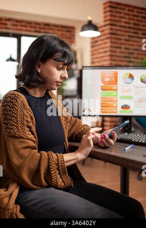 Geschäftsfrau, die E-Mails auf dem Smartphone liest, während ein Desktop-Monitor Geschäftsanalysen am Schreibtisch anzeigt. Weibliche Mitarbeiterin, die ein mobiles Gerät hält, um mit Familie und Freunden in einem Raum mit gemauerten Wänden zu chatten. Stockfoto