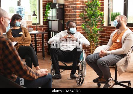 afroamerikanischer männlicher Rollstuhlfahrer, der während der Therapiesitzung mit der Gruppe spricht. Männliche und weibliche Patienten hören aufmerksam auf schwarze Männer und bieten Unterstützung durch die Gemeinschaft beim Programm für psychische Gesundheit. Stockfoto