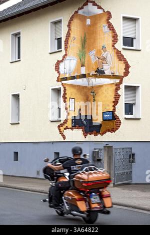 Haus mit lustiger Wandmalerei, Blick in die Innenräume, Mann liest Zeitung auf Toilette, Frau im Wohnzimmer beobachtet den Tatort im Fernsehen, Motorradfahrer, Fahrrad Stockfoto
