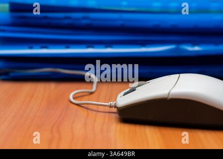 Eine Computermaus sitzt neben einem Stapel blauer Akten auf einem hölzernen Schreibtisch. Der Arbeitsbereich ist organisiert und kann während der Bürozeiten produktiv arbeiten. Stockfoto