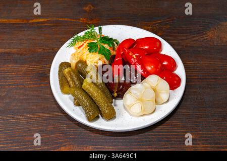 Eine lebendige Vielfalt an eingelegtem Gemüse, einschließlich Gurken, Kirschtomaten und Knoblauch, ordentlich auf einem weißen Teller angeordnet. Frische Kräuter sorgen für einen Hauch o Stockfoto