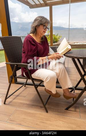 Vertikale Aufnahme einer Latina in ihren 70ern, die eine Brille trägt und ein Buch liest, die draußen an einem Rattan-Tisch sitzt Stockfoto