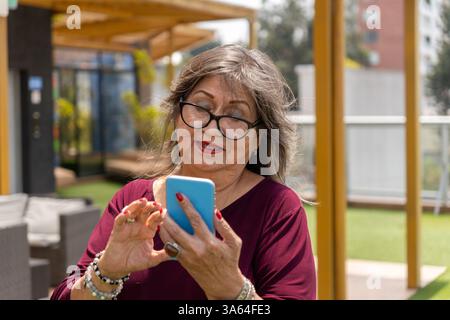 Porträt einer älteren Frau mit Brille, die morgens draußen ihr Handy benutzt Stockfoto