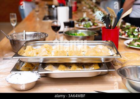 Ein Tablett mit frisch zubereiteten Ravioli, mit Mehl bestäubt, steht auf einer Küchentheke, umgeben von Zubereitungswerkzeugen und Zutaten. Stockfoto