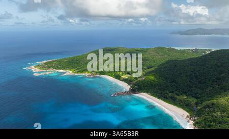 Türkisfarbene Meereswellen treffen auf eine felsige Küste, die von grünen bewaldeten Hügeln begrenzt wird. La Digue, Seychellen. Stockfoto