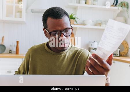 Afroamerikaner mit Brille, die Belege sorgfältig überprüft, mit Daten auf seinem Laptop verglichen, Haushaltsfinanzen verwaltet und ein Budget in seiner modernen Küche erstellt Stockfoto