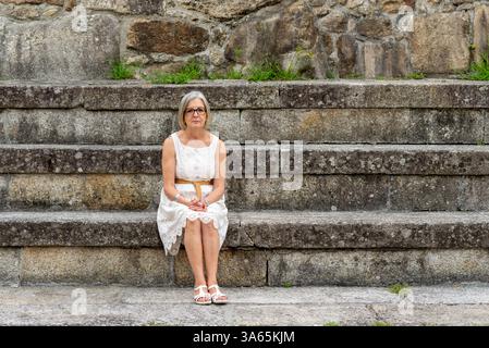 Porträt einer seriösen Seniorin, die sich entspannt und posiert, während sie auf Steintreppen vor einer alten Steinmauer sitzt Stockfoto