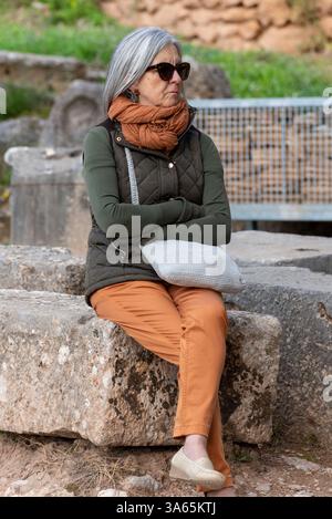 Eine reife Frau in Herbstkleidung sitzt draußen auf einem Stein und reflektiert. Ihr Schal, ihre gesteppte Jacke und ihre Sonnenbrille zeigen eine stilvolle und doch entspannte Note Stockfoto