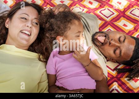 Lachende, vielfältige Familie, die sich auf einer gemusterten Picknickdecke im Park entspannt und einen verspielten Moment im Freien genießt. Stockfoto
