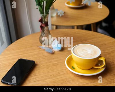 Gelbe Kaffeetasse sitzt auf einem Tisch neben einem Handy. Die Tasse ist mit Kaffee gefüllt und hat ein weißes Design. Der Tisch ist aus Holz und hat eine Vase mit Blumen darauf. Die Szene ist gemütlich Stockfoto