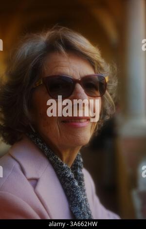 Ältere Frau mit Sonnenbrille, warm gekleidet mit Schal und Mantel, sanft lächelnd. Hintergrund weist auf eine städtische oder architektonische Umgebung hin Stockfoto