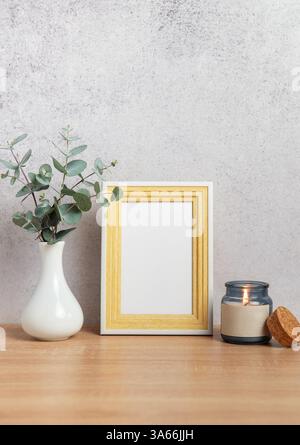 Holzrahmenmockup mit brennender Kerze und Eukalyptuszweigen in Vase auf Holztisch vor grauer Wand Stockfoto