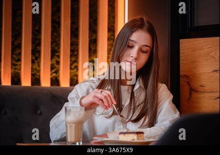 Eine junge Frau in einem weißen Hemd genießt ein Stück Kuchen und einen Latte in einem stilvollen Café. Warme Beleuchtung und modernes Deko schaffen eine gemütliche und einladende Atmosphäre Stockfoto