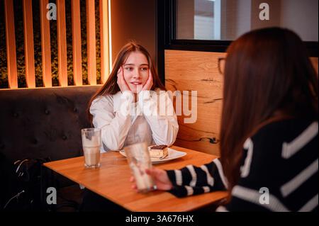 Zwei junge Frauen sitzen an einem Café-Tisch und teilen sich ein warmes und fesselndes Gespräch. Eine lächelt mit Händen auf ihren Wangen, während die andere zuhört. Gemütliches Ambi Stockfoto