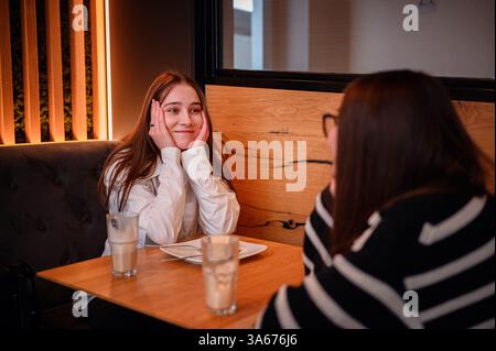 Zwei junge Frauen sitzen an einem Café-Tisch und teilen sich ein warmes und fesselndes Gespräch. Eine lächelt mit Händen auf ihren Wangen, während die andere zuhört. Gemütliches Ambi Stockfoto