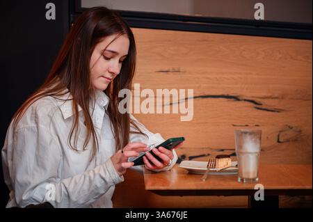 Eine junge Frau in einem weißen Hemd sitzt an einem Café-Tisch und konzentriert sich auf ihr Smartphone. Ein Latte und ein teilweise gegessen Dessert stehen auf dem Tisch, was einen Coz schafft Stockfoto