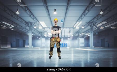 Lächelnder Bauarbeiter, der mit überkreuzten Armen in einem großen leeren Lager steht Stockfoto