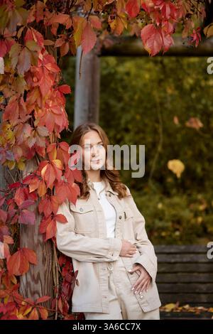 Herbstporträt: Stilvolle junge Frau in Beige Outfit posiert von Holzpergola mit roten Herbstblättern. Gemütliches Herbstporträt in landschaftlich reizvollem Garten Stockfoto