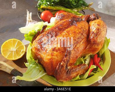 Ein perfekt geröstetes Huhn sitzt auf einem Bett aus knusprigem Salat, umgeben von lebendigem Gemüse und einer Zitrone, bereit für einen Familientreffen. Stockfoto