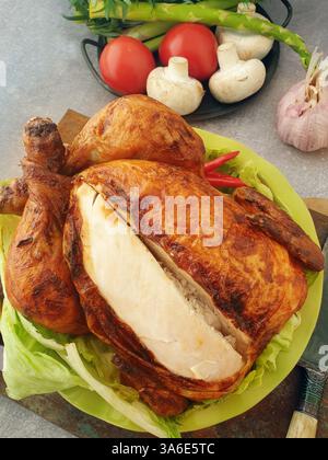 Ein perfekt geröstetes Huhn sitzt auf einem Bett aus knusprigem Salat, umgeben von lebendigem Gemüse und einer Zitrone, bereit für einen Familientreffen. Stockfoto