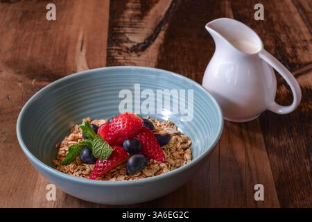 Blaue Schüssel Frühstücksgranola mit frischen Erdbeeren, Heidelbeeren und kleinem weißen Keramikkrug mit Joghurt oder Milch auf rustikalem Holztisch Stockfoto