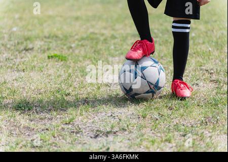 Nahaufnahme von Kinderbeinen in schwarzen Socken und roten Stollen, die auf dem Grasfeld stehen und einen Fuß auf Fußball setzen. Sonniger Tag, heller Schatten auf trockenem Gras, Fokus o Stockfoto