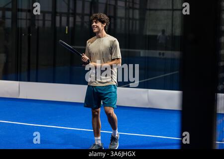Junger Mann, der Padel-Tennis auf dem Hallenplatz spielt, lächelt und Schläger hält Stockfoto
