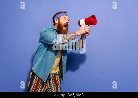 Stilvoller Mann mit Bart, der durch Megaphon schreiend in lässigem Outfit auf leuchtendem violettem Hintergrund Stockfoto