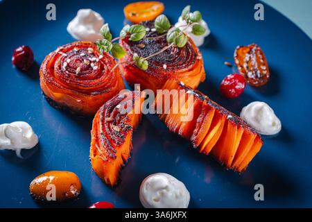 Karotten-Dessert aus dünnen Scheiben, die zu einem Brötchen zusammengelegt werden, serviert mit Kumquat, Preiselbeeren und Sahne auf einem blauen Teller. Nahaufnahme. Hochwertige Fotos Stockfoto