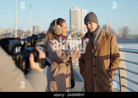 Junger Mann und Frau in beigefarbenen Mänteln, die vor einer Videokamera stehen, die ein Außeninterview am Flussufer aufnehmen Stockfoto
