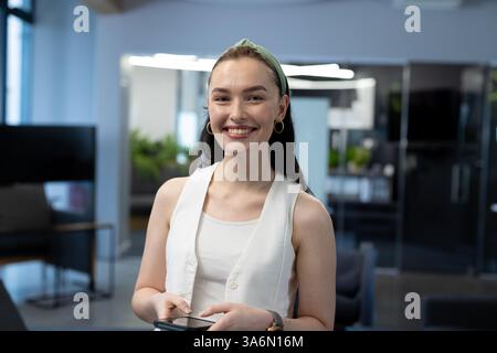 Das Smartphone im Büro halten, eine lächelnde Frau, die selbstbewusst und zugänglich aussieht Stockfoto