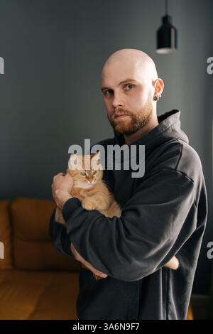Vertikales Porträt eines hübschen Glatzenmannes, der sich um eine Ingwerkatze dreht und in die Kamera schaut. Bärtiger Tierbesitzer in schwarzem Hoodie, der eine katzenartige Begleiterin umschließt Stockfoto