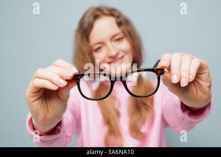 Die lächelnde Frau in Rosa hält eine stilvolle Brille in Richtung der Kamera und erzeugt einen perspektivischen Effekt vor einem pastellblauen Hintergrund. Stockfoto
