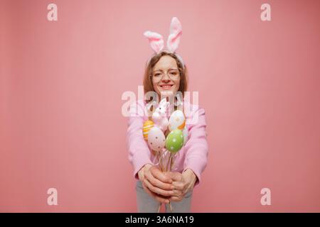 Glückliche Frau in Hasenohren, die einen Strauß bunter Ostereier hält und verspielt lächelt. Festliche Frühlingsstimmung, Urlaubsfreude und saisonale Feierlichkeiten Stockfoto
