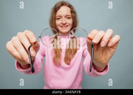 Eine lächelnde Frau in einem rosafarbenen Pullover hält eine runde Brille vor einem hellblauen Hintergrund in Richtung Kamera. Stockfoto