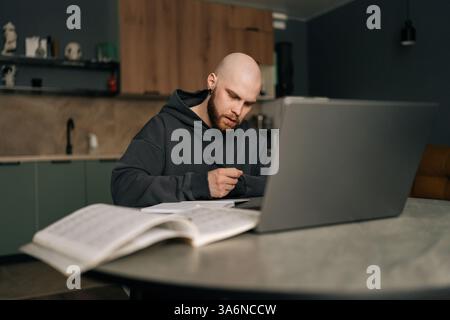 Bärtiger, kahlköpfiger junger Mann im Hoodie, der sich Notizen macht, am Küchentisch mit Laptop sitzt, Online-Geschäfte managt und den Komfort der Arbeit genießt Stockfoto