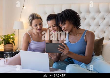 Vielfältige Freundinnen, die sich im Bett entspannen, Tablet und Laptop nutzen und gemeinsame Freizeit genießen Stockfoto