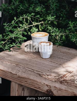Ein flaches weiß und ein Kaffee-Macchiato in schönen Keramikbechern auf einem Holztisch mit Pflanzen im Hintergrund Stockfoto