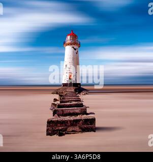 Alter weißer Leuchtturm mit roter Spitze und einem Betonblockweg, der zu seiner roten Eingangstür am abstrakten Strand mit einem hellblau-weißen Himmel führt Stockfoto