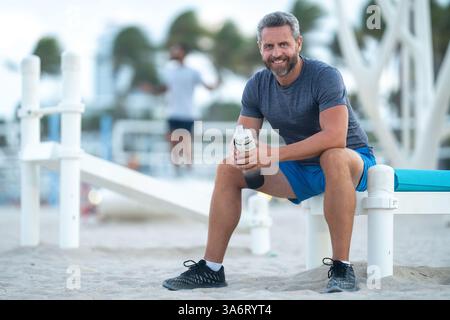 Sport-Fitness-Mann, der draußen auf einer Bank ruht. Reifes Fitnesssportmodell, das nach dem Training auf einer Bank sitzt. Ein Sportmann macht nach dem Fitnesstraining eine Pause Stockfoto