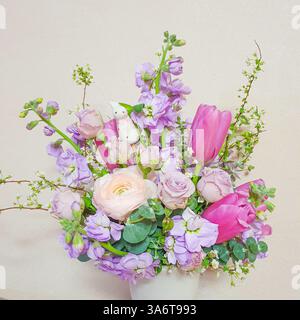 Blumenarrangement in einer weißen Vase mit rosa Tulpe, roter Rose, lila Brühe und grünem Laub. Kleine weiße Kaninchenfigur. Frühlingsatmosphäre Stockfoto