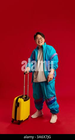 Ganzkörperfoto eines glücklichen, lächelnden jungen asiatischen Mannes, der mit Gepäck posiert, bereit für die Reise vor rotem Hintergrund. Stockfoto