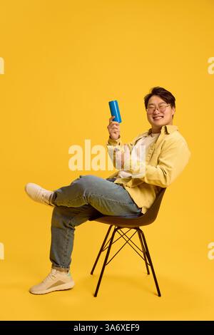 Glücklicher junger asiatischer Mann, der auf einem Stuhl sitzt und den Daumen nach oben zeigt, mit leerem Platz vor sonnigem gelbem Hintergrund. Stockfoto