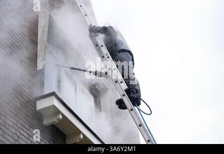 Mann im wasserdichten Anzug reinigt schmutzige Ziegelfassade des Wohnhauses mit Hochdruckwasserstrahl. Hochdruckreinigung der Fassade Stockfoto