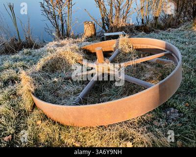 Ein verwittertes gusseisernes Mühlenrad liegt in frostigem Gras bei Bruchmühle, bedeckt mit Eiskristallen an einem kalten Wintermorgen. Stockfoto