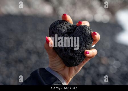 Menschliche Hand der Frau mit rosafarbenen Nägeln, die schwarzen vulkanischen Stein in Herzform unter schwarzem Sandhintergrund in hohem Kontrast halten Stockfoto